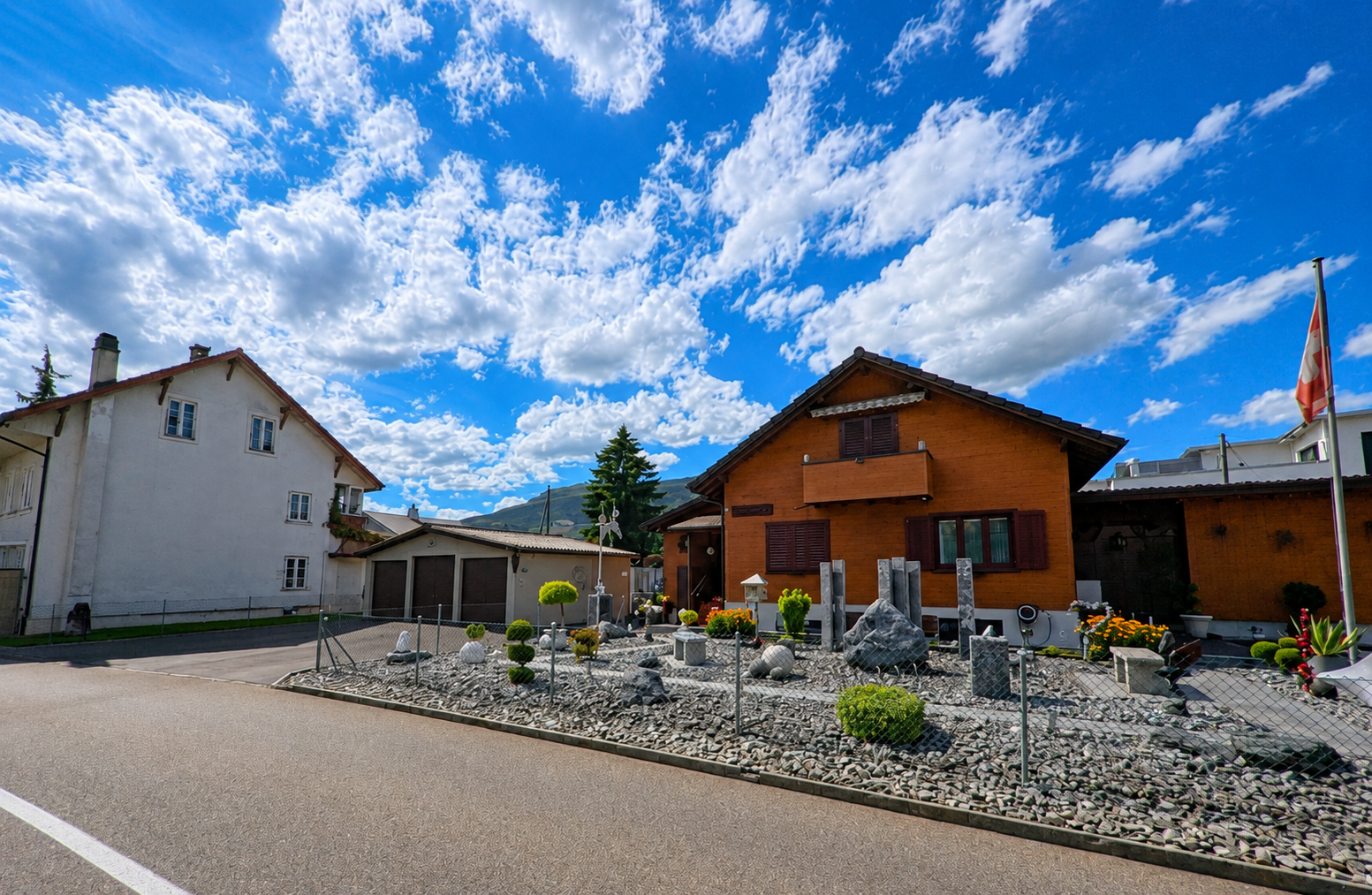 In Niederbuchsiten (4626, Kanton Solothurn) stehen Wohnhäuser mit gepflegtem Steingarten entlang einer ruhigen Dorfstrasse unter blauem Himmel mit Wolken.