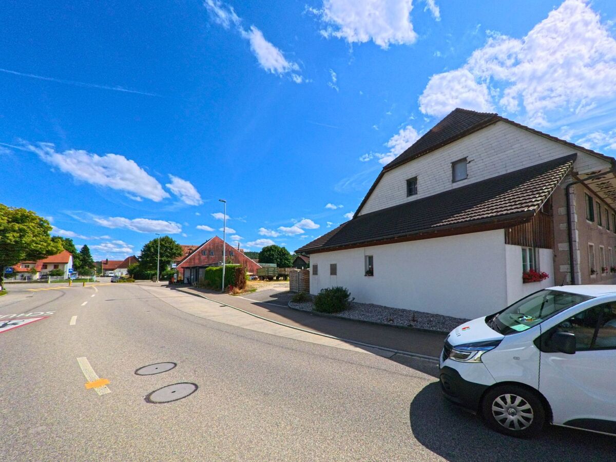 Entlang der Hauptstrasse von Niederbuchsiten (4626, Kanton Solothurn) reihen sich Wohn- und Bauernhäuser mit Vorgärten, begleitet von einem vorbeifahrenden Lieferfahrzeug.
