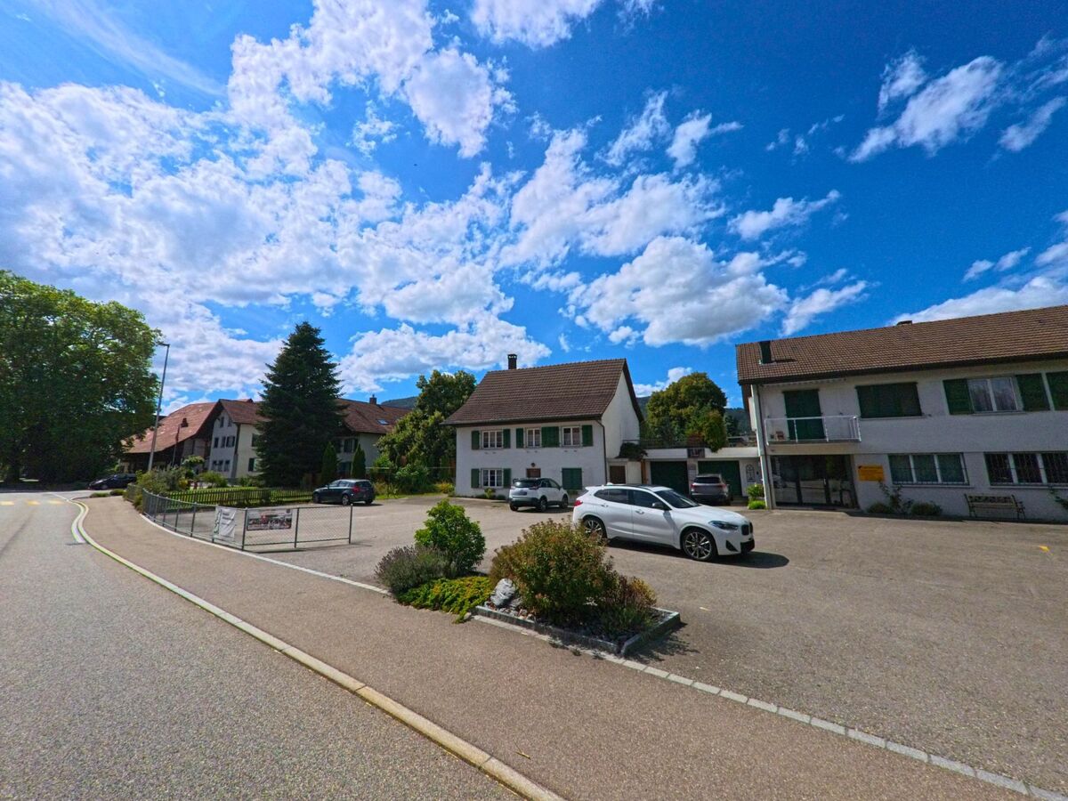 In Niederbuchsiten (4626, Kanton Solothurn) erstreckt sich eine Strasse entlang eines offenen Parkplatzes mit angrenzenden Wohnhaeusern und einem kleinen Geschäftshaus.