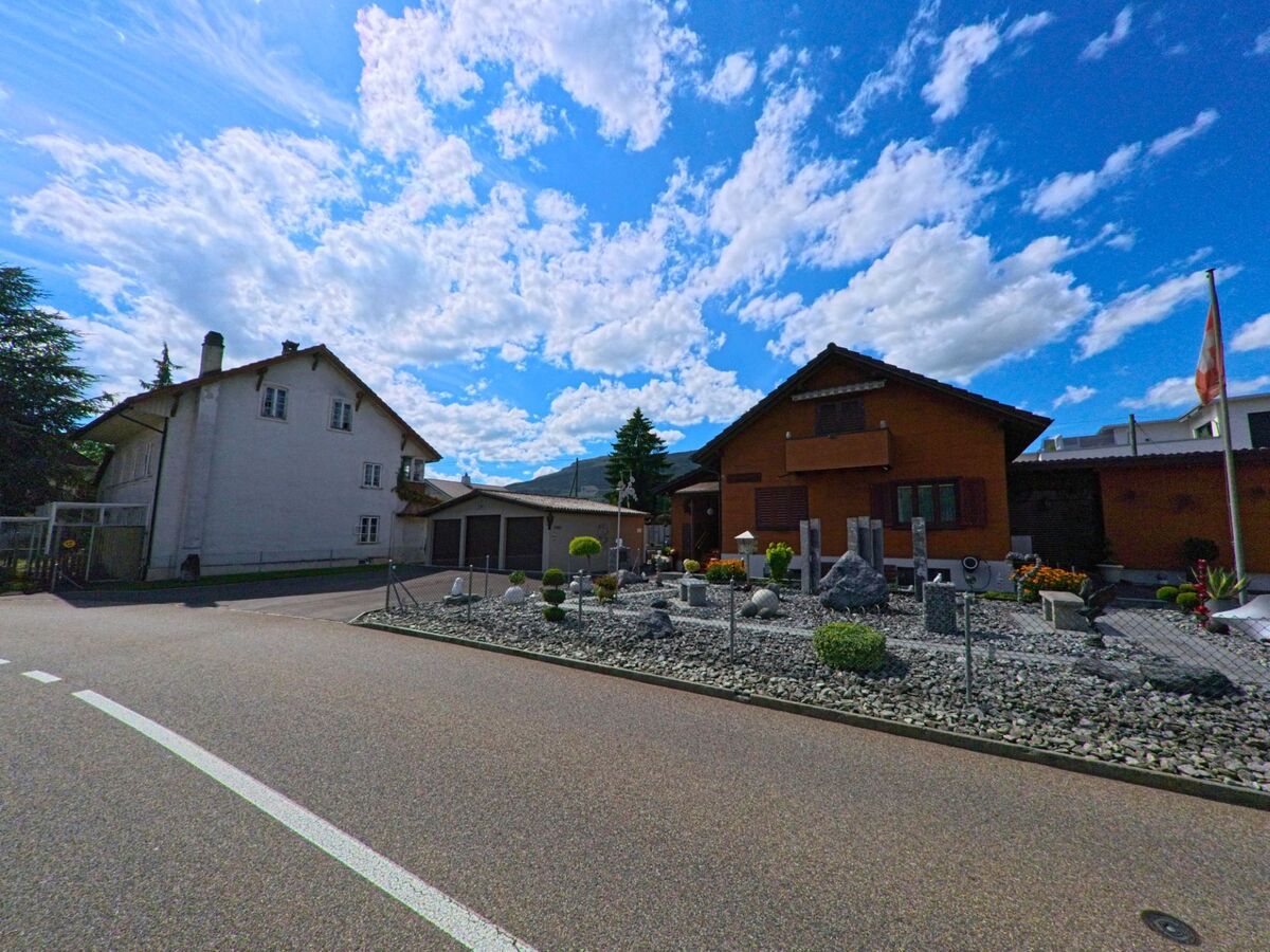 An einer Strasse in Niederbuchsiten (4626, Kanton Solothurn) steht ein Holzhaus mit dekorativem Steingarten, daneben ein weisses Wohnhaus mit Garagen.