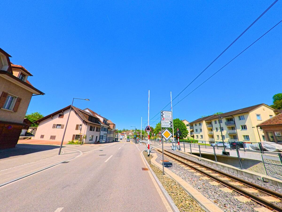 In Niederdorf (4435, Kanton Baselland) kreuzt die Bahnlinie an einem beschrankten Übergang die Hauptstrasse, flankiert von Wohnhäusern und Wegweisern zu benachbarten Gemeinden.