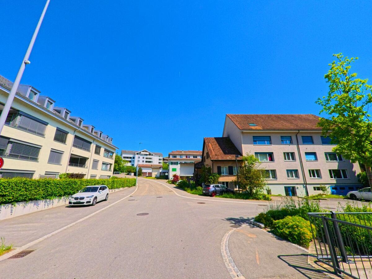 In Niederdorf (4435, Kanton Baselland) steigt eine Strasse zwischen modernen Mehrfamilienhäusern sanft an, begleitet von geparkten Autos und begrünten Vorgärten.