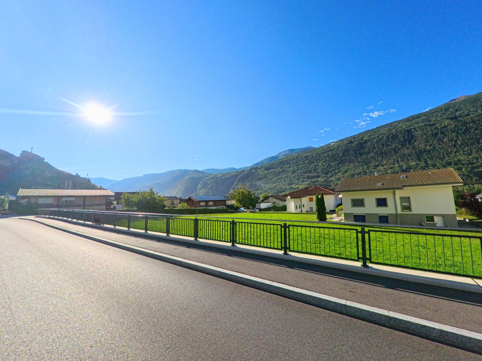 Sonnige Strasse mit grüner Wiese und Wohnhäusern in Niedergesteln (3942, Kanton Wallis), umgeben von bewaldeten Bergen.