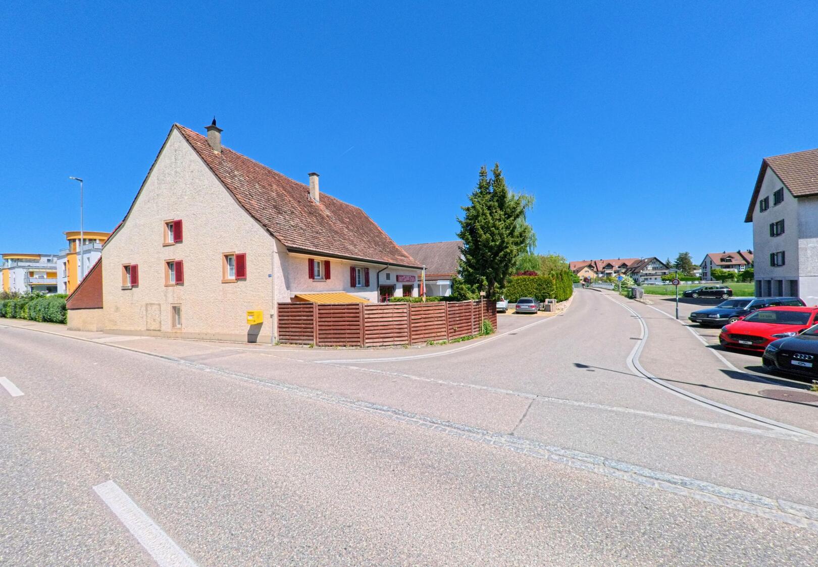 An einer Hauptstrasse in Niederglatt (8172, Kanton Zürich) steht ein traditionelles Wohnhaus mit rotem Fensterladen neben modernen Gebäuden unter klarem, blauem Himmel.