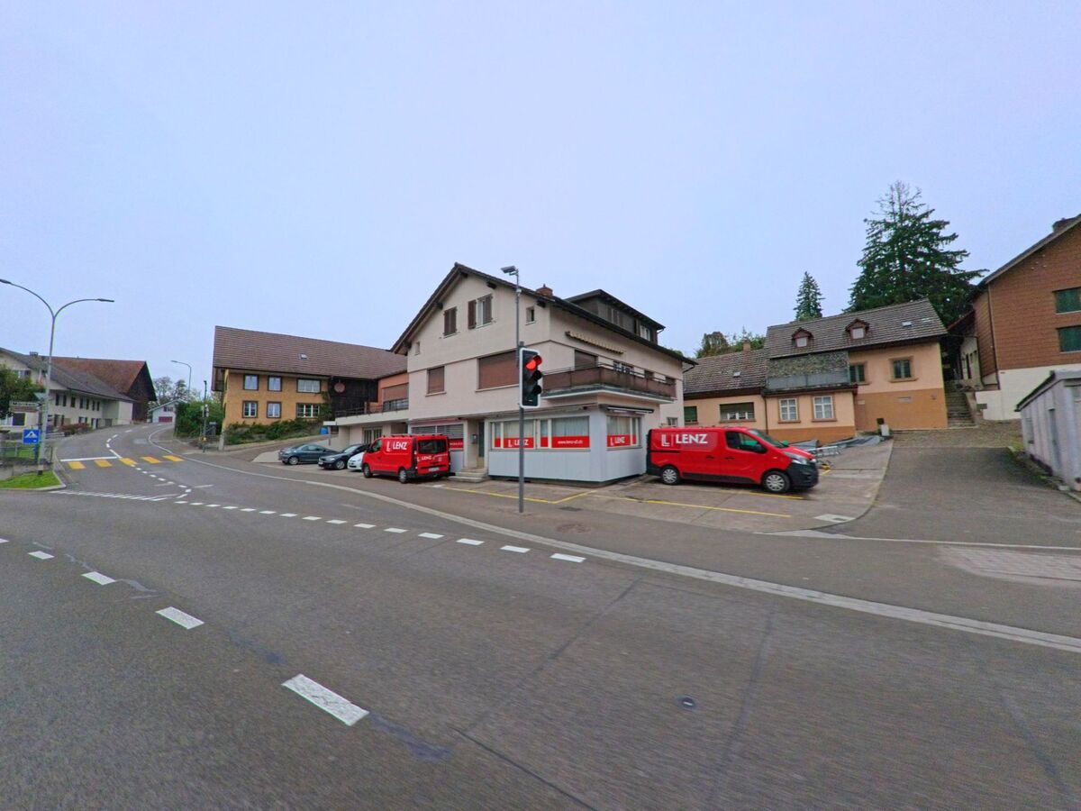 An einer Strassenkreuzung in Niederlenz (5702, Kanton Aargau) stehen mehrere Gebaeude mit roten Lieferwagen der Firma Lenz vor einer Ampel.