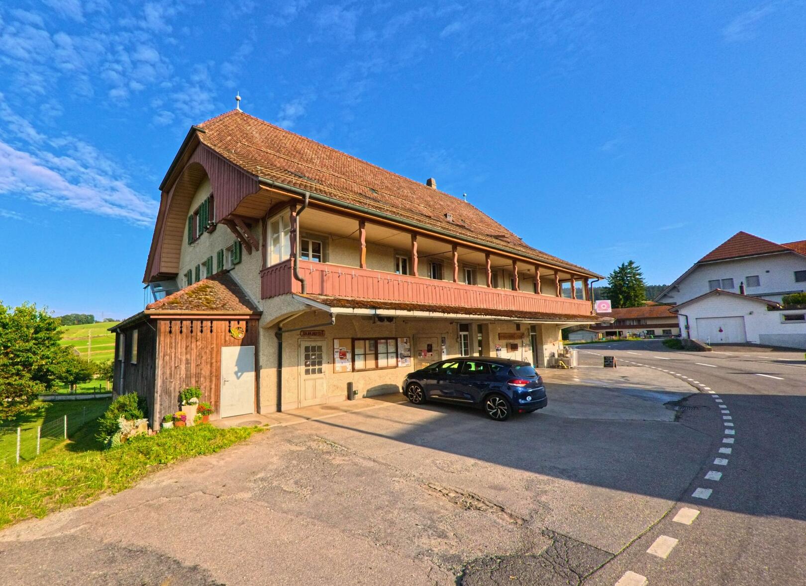 Ein traditionelles Gebaeude mit Holzveranda und rotem Ziegeldach steht in Niedermuhlern (3087, Kanton Bern) an einer leicht gekruemmten Dorfstrasse.
