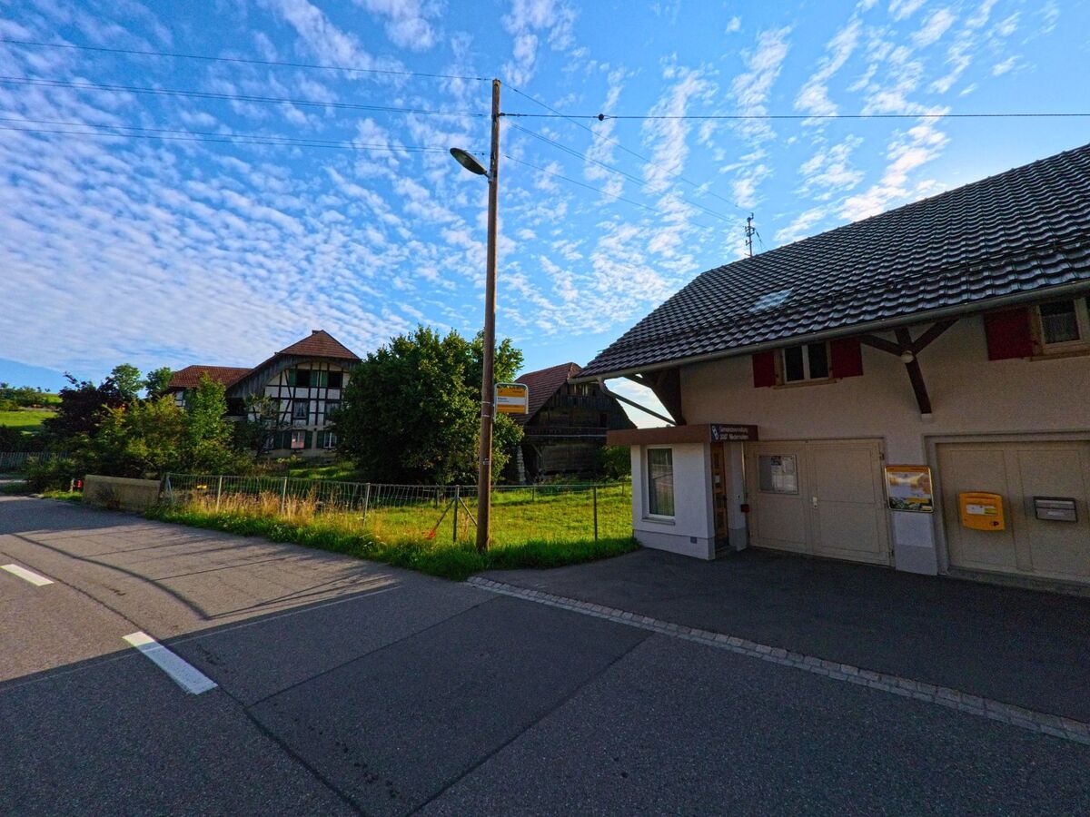 Ein kleines Gebaeude mit Briefkasten und Bushaltestelle steht in Niedermuhlern (3087, Kanton Bern) gegenueber eines traditionellen Fachwerkhauses.