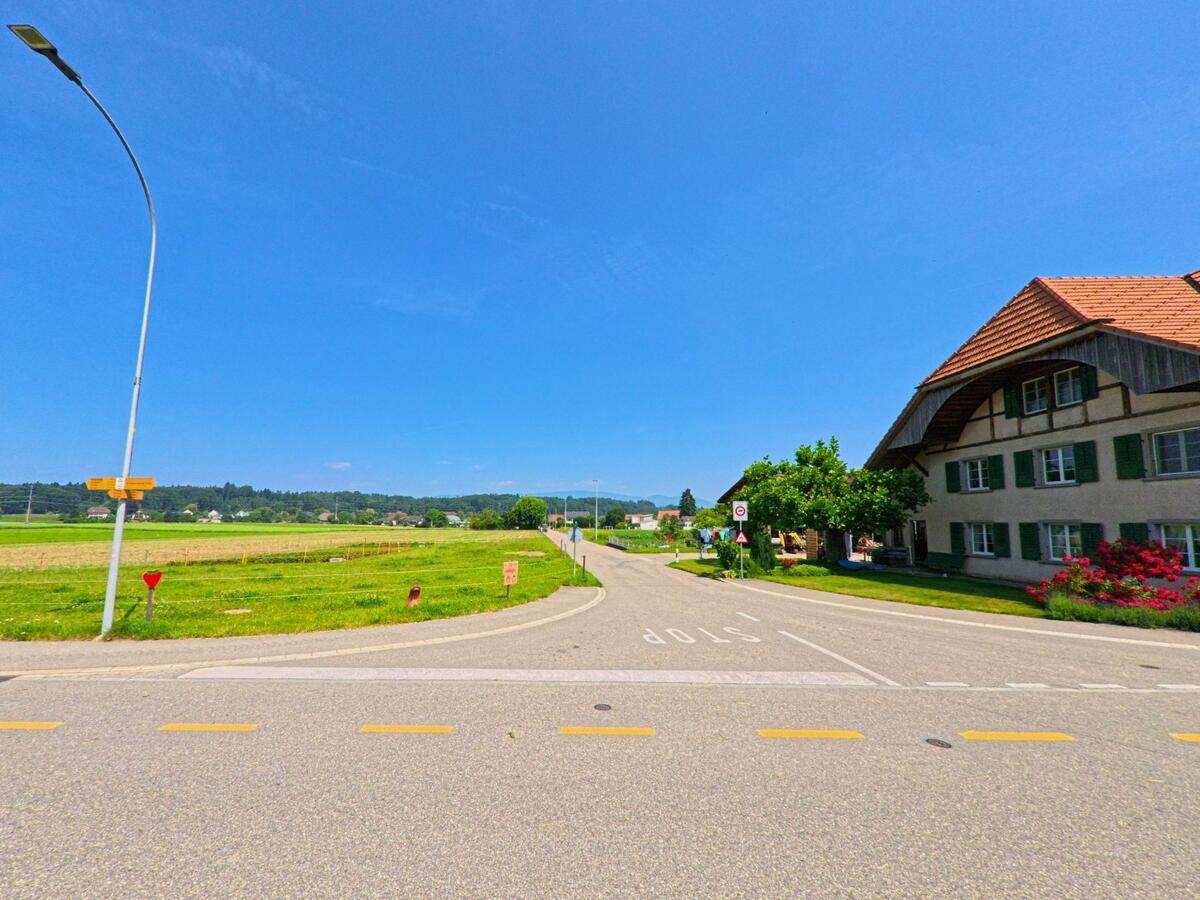 Am Rand von Niederönz (3362, Kanton Bern) führt eine schmale Strasse an einem traditionellen Bauernhaus mit grünen Fensterläden vorbei in eine weite Ebene mit Feldern.