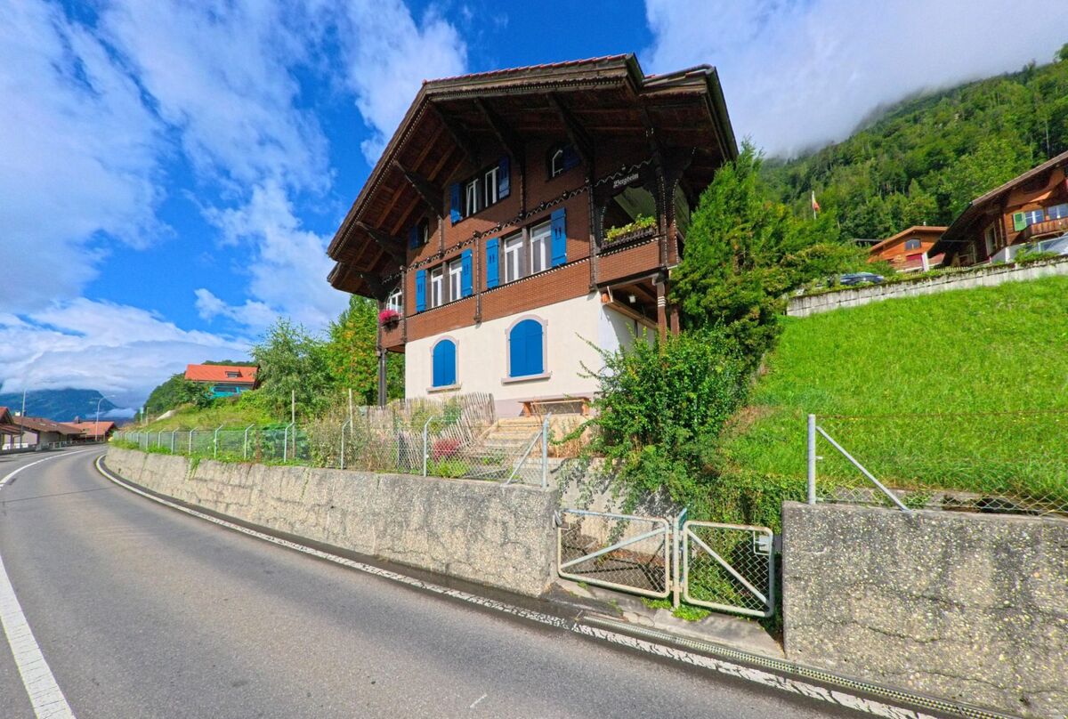 In Niederried bei Interlaken (3853, Kanton Bern) steht ein traditionelles Chalet mit blauen Fensterläden direkt an einer kurvigen Bergstrasse.