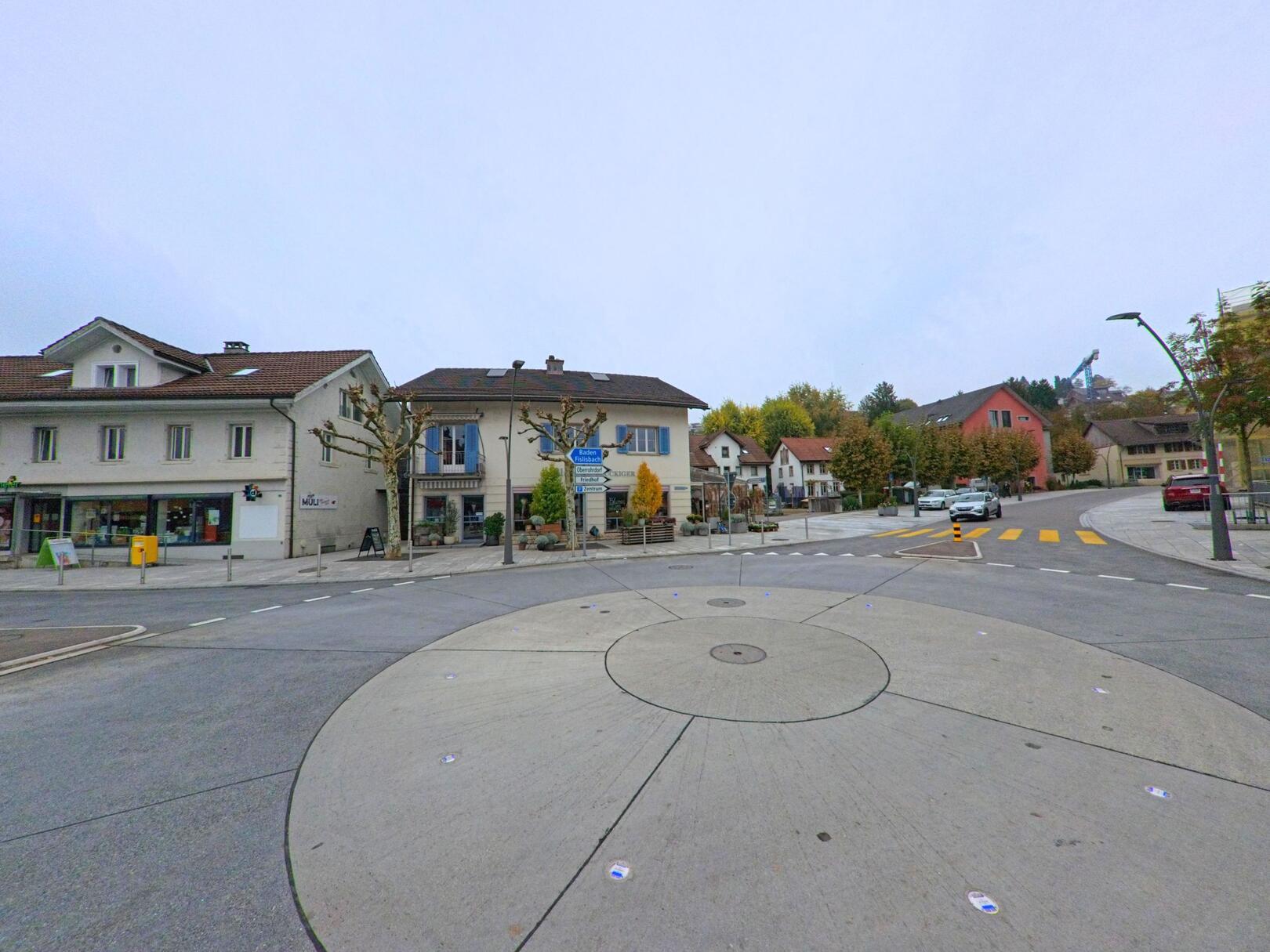 Im Zentrum von Niederrohrdorf (5443, Kanton Aargau) liegt ein kleiner Kreisverkehr, umgeben von Wohn- und Geschaeftshaeusern mit herbstlich gefaerbten Baeumen.