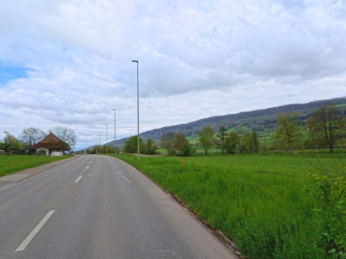 In Niederweningen, PLZ 8166 im Kanton Zürich, erstreckt sich eine lange Landstrasse durch offene Felder mit Blick auf bewaldete Hügel.