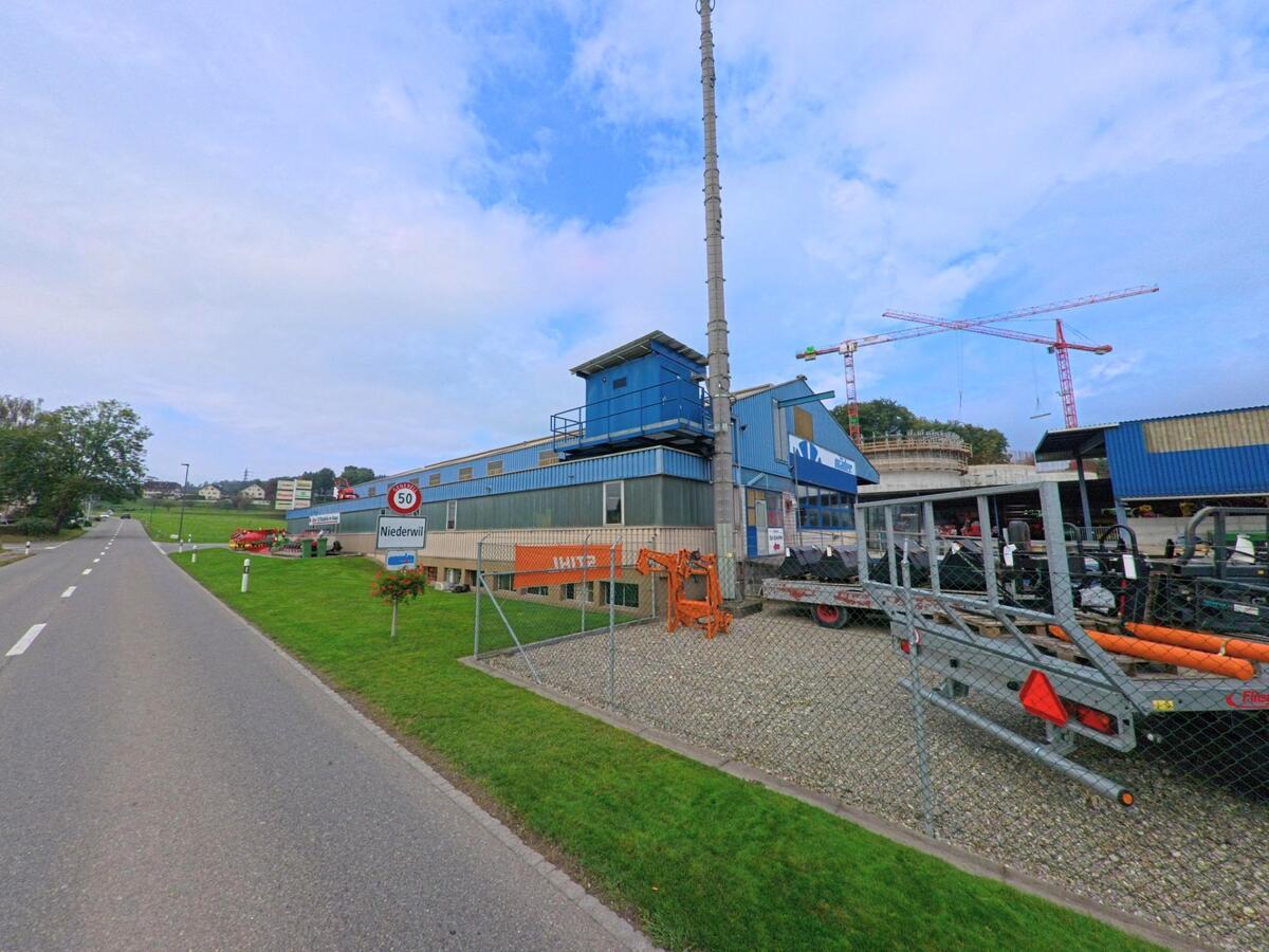 Am Ortseingang von Niederwil (5524, Kanton Aargau) steht ein Gewerbebetrieb mit Baugeraeten und Kranen, waehrend die Landstrasse in Richtung Dorf fuehrt.