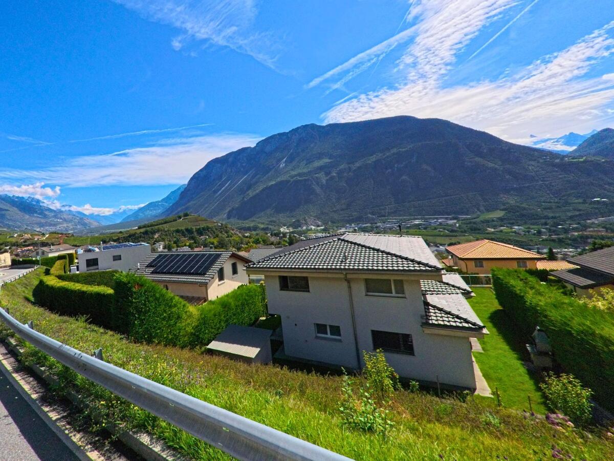 Von einer Anhöhe in Noble-Contrée (3968, Kanton Wallis) blickt man auf moderne Einfamilienhäuser mit Gärten und die umliegenden Bergketten.
