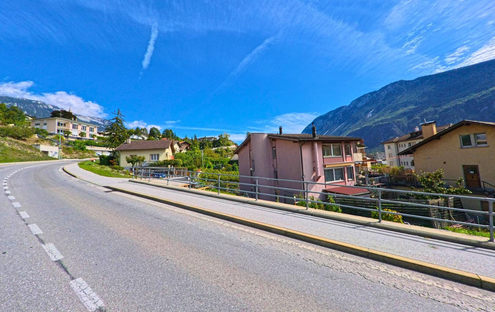 Eine kurvige Strasse führt in Noble-Contrée (3968, Kanton Wallis) an Wohnhäusern vorbei, während im Hintergrund die Berghänge sichtbar sind.