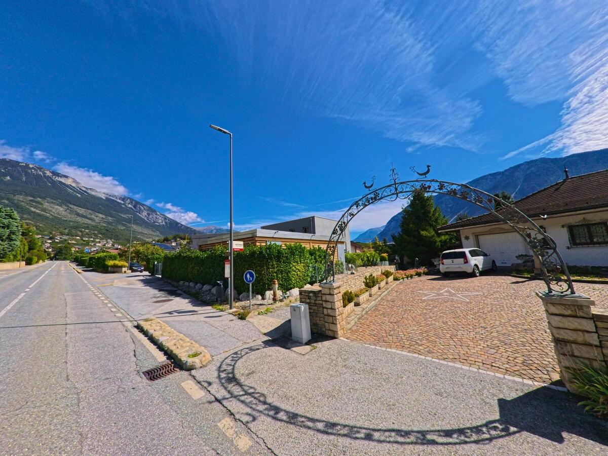 Ein verzierter Torbogen führt in Noble-Contrée (3968, Kanton Wallis) zu einem gepflasterten Hof mit geparktem Auto, während im Hintergrund die Berge sichtbar sind.