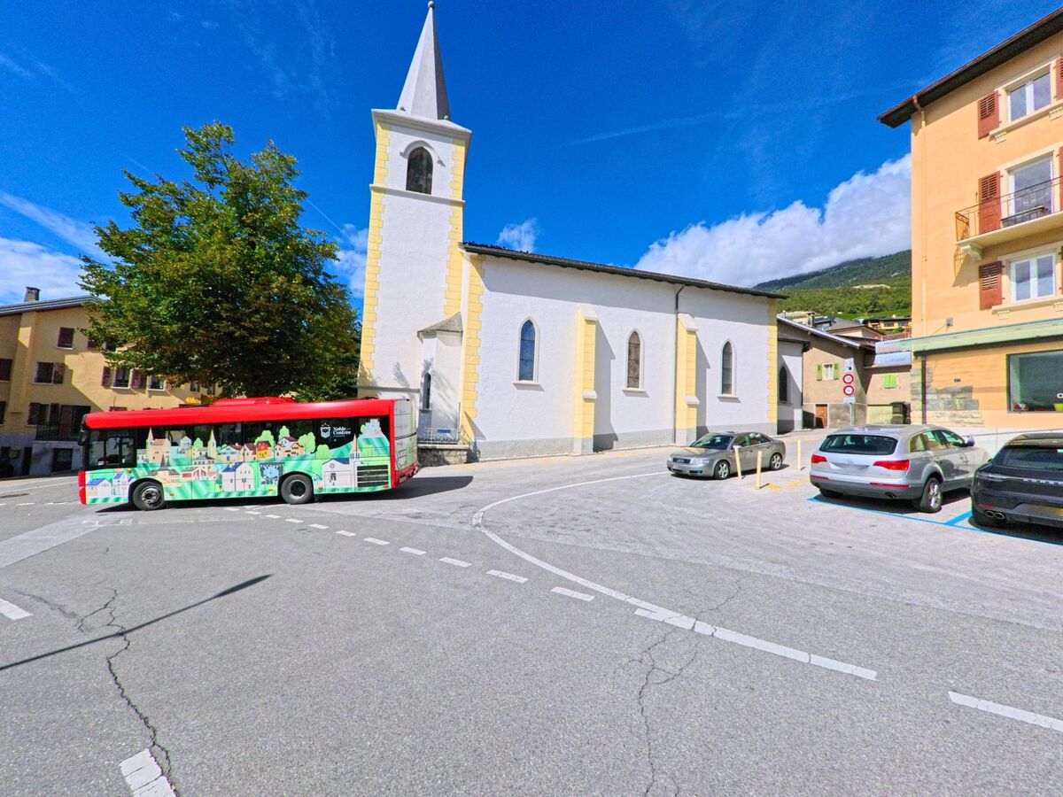 Vor der Kirche mit gelben Ecksteinen in Noble-Contree (3972, Kanton Wallis) faehrt ein bunter Bus vorbei, waehrend Autos daneben parken.