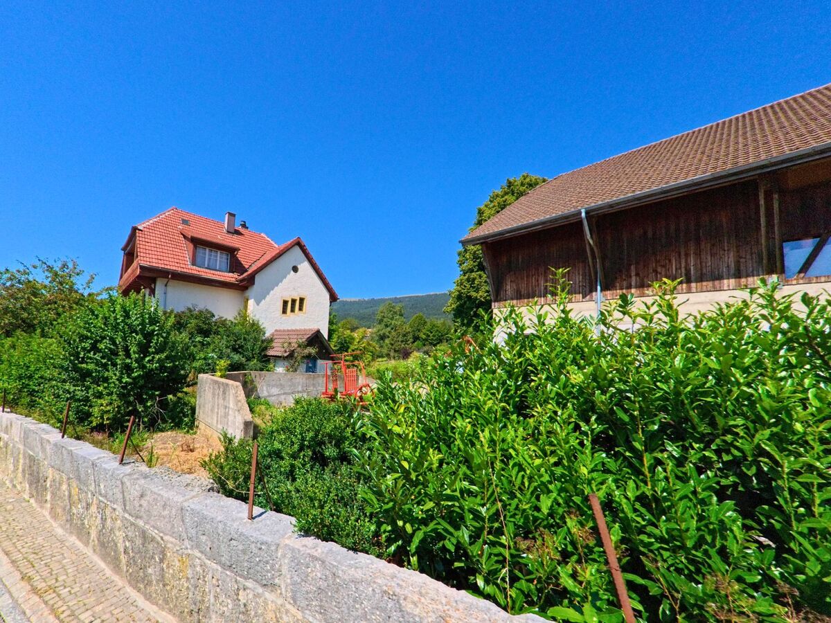 Zwischen dichten Bueschen in Nods (2518, Kanton Bern) stehen ein Wohnhaus mit rotem Ziegeldach und eine angrenzende Scheune unter klarem Himmel.