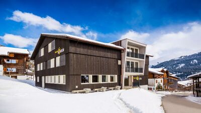 Die Nova Casa Spinatscha in Tujetsch (7188, Kanton Graubuenden) steht mit moderner Fassade und Holzelementen in einer verschneiten Berglandschaft.