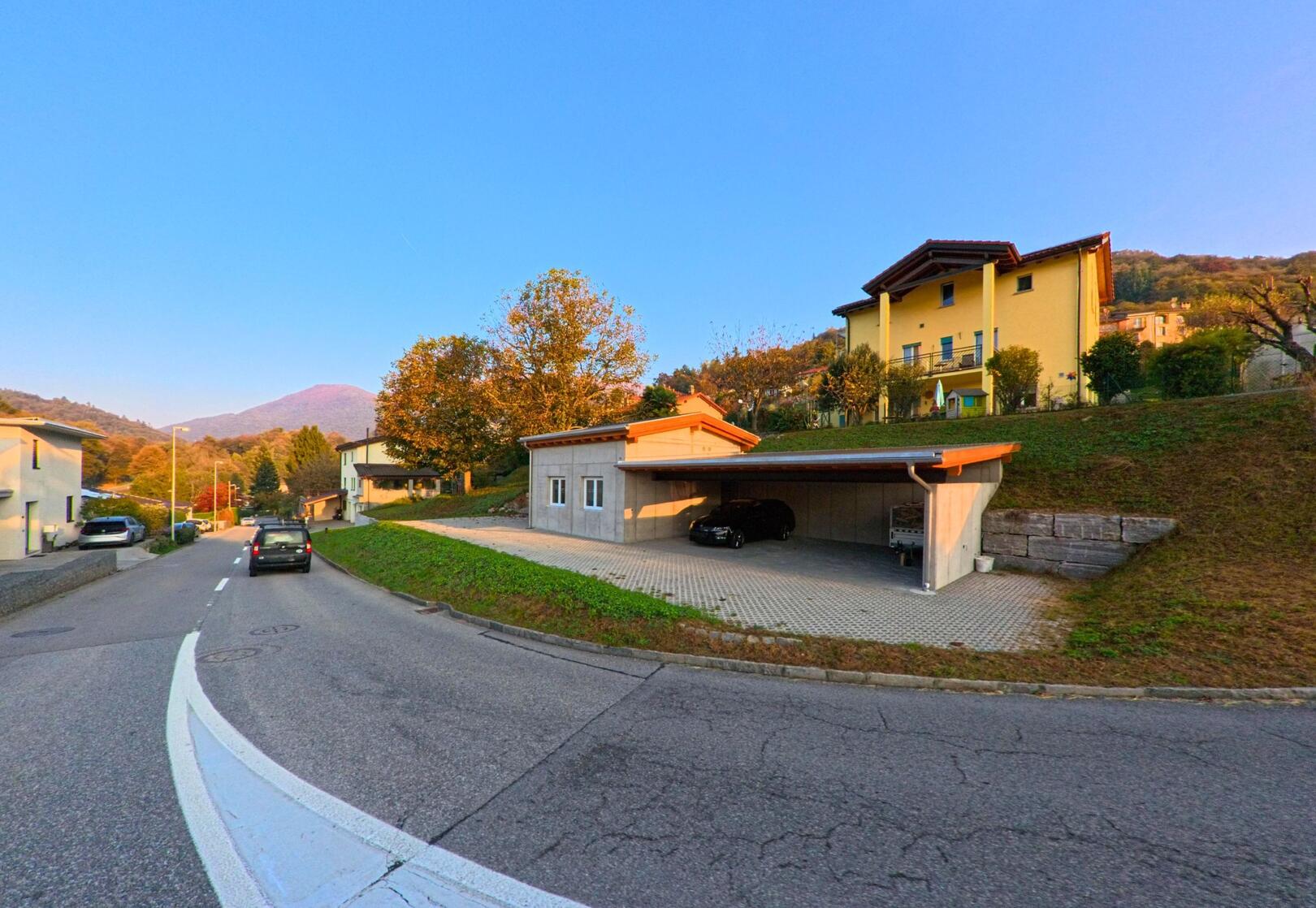 Entlang einer ruhigen Strasse zeigt sich in Novaggio (6986, Kanton Tessin) ein Wohnbereich mit Einfamilienhäusern, Carport und Hanglage.