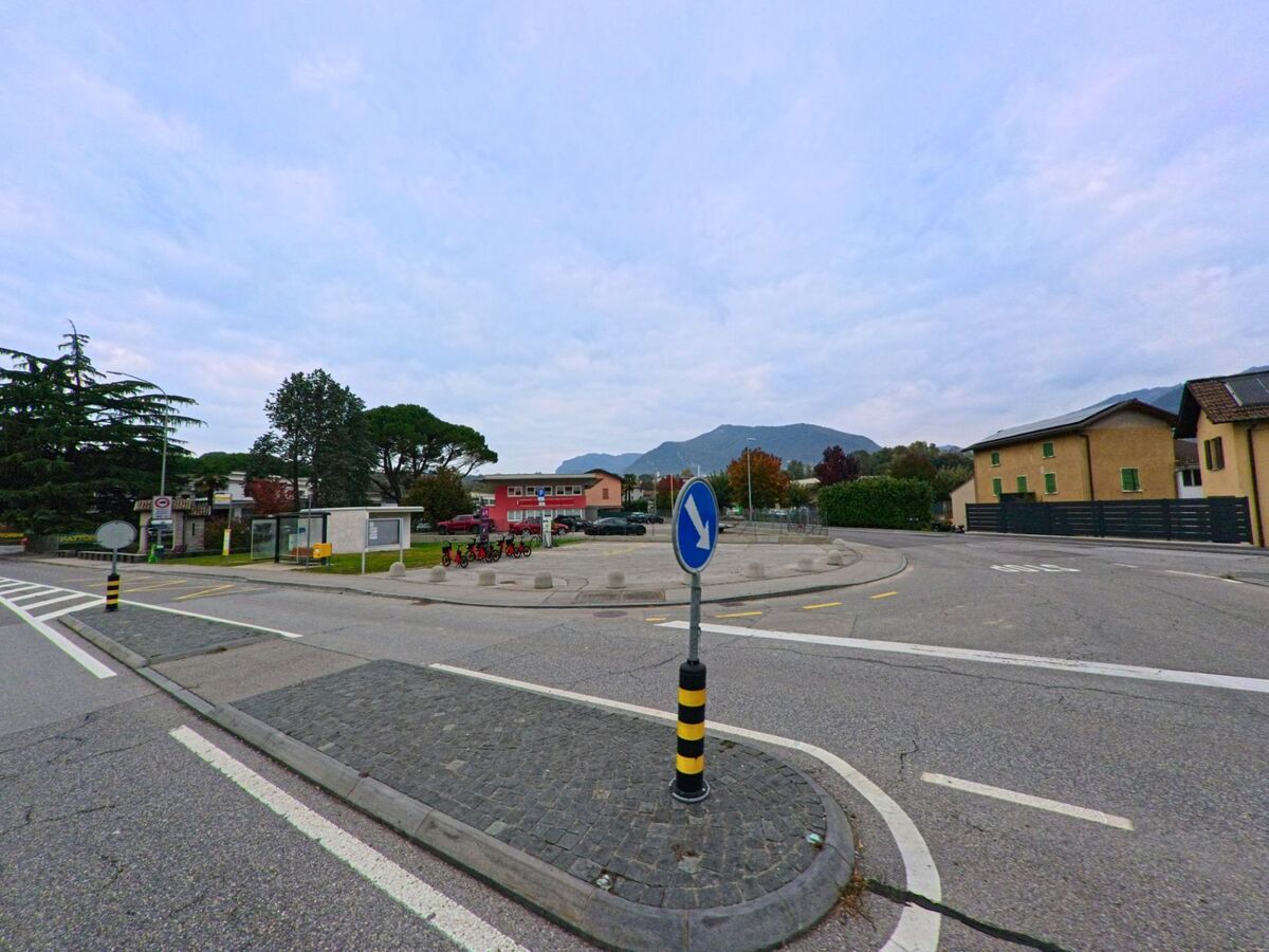 Ein kleiner Verkehrsknotenpunkt mit Radverleihstation und Bushaltestelle liegt in Novazzano (6883, Kanton Tessin) vor einer Kulisse aus Huegeln und Wohnhaeusern.