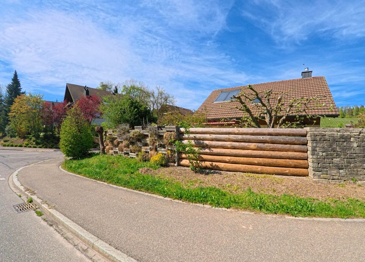 In Nürensdorf, PLZ 8309 im Kanton Zürich, schützt eine Gartenmauer aus Holzstämmen und Stein ein Einfamilienhaus mit Ziegeldach an einer ruhigen Kurve.
