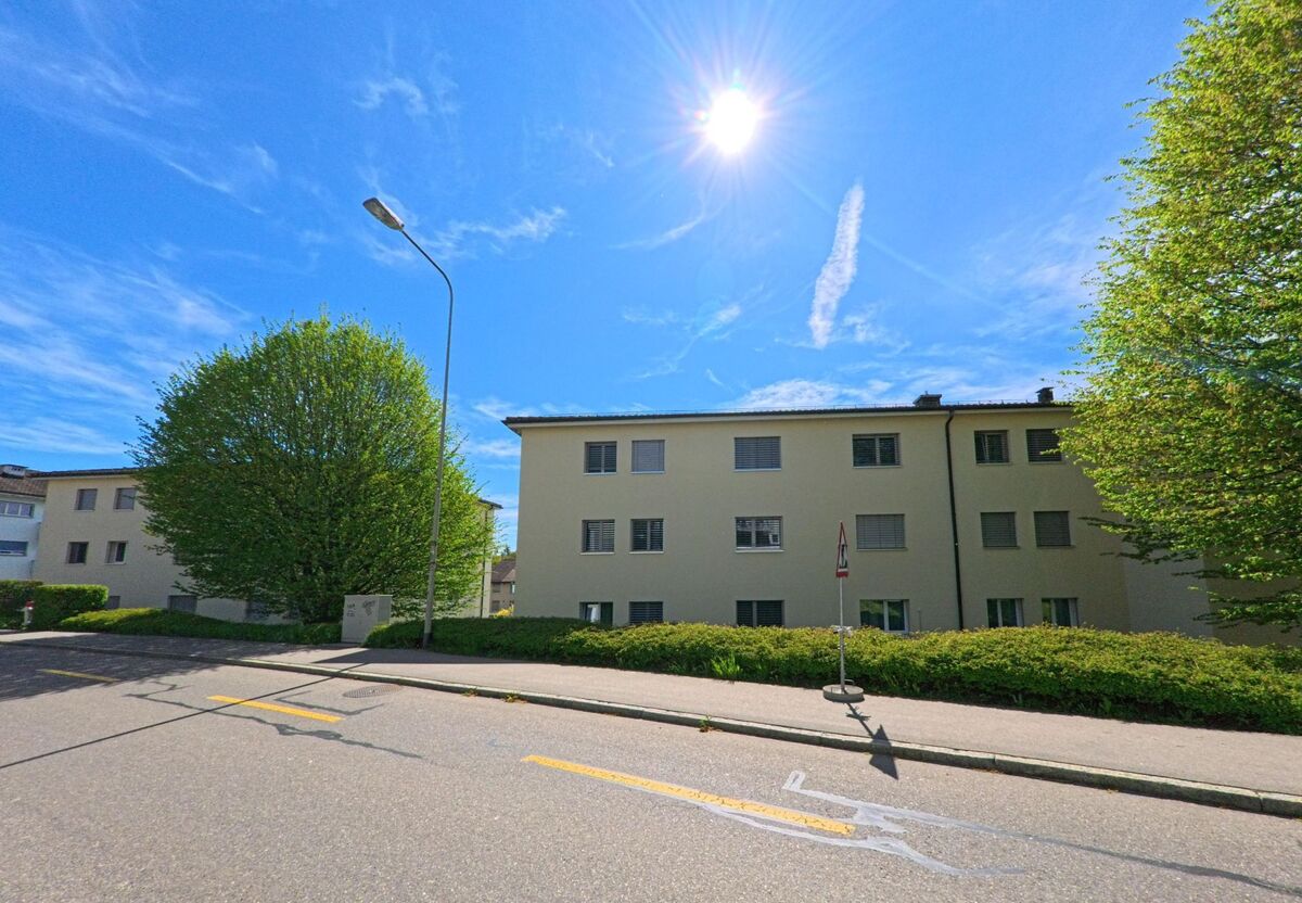 In Nürensdorf, PLZ 8309 im Kanton Zürich, stehen schlichte Mehrfamilienhäuser entlang einer sonnigen Quartierstrasse mit gepflegtem Gehweg.