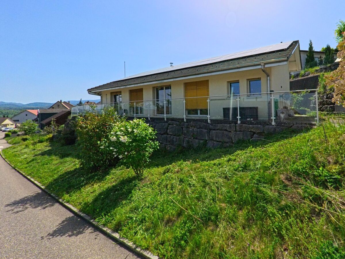 Ein modernes Einfamilienhaus mit Terrasse und Glasgelaender erhebt sich in Nuglar-St. Pantaleon (4412, Kanton Solothurn) ueber einer kleinen Gruenflaeche am Hang.