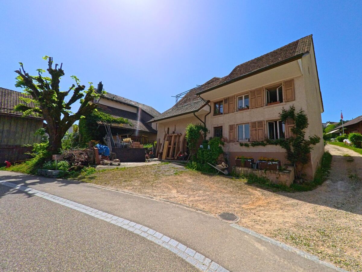 Ein traditionelles Wohnhaus mit Holzlaeden und begruenter Fassade steht in Nuglar-St. Pantaleon (4412, Kanton Solothurn) neben einem Hof mit einem markant beschnittenen Baum.
