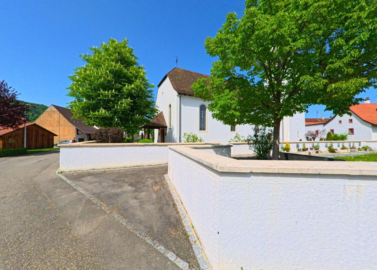 Die Kirche von Nuglar-St. Pantaleon (4421, Kanton Solothurn) liegt hinter einer weissen Mauer, umgeben von gruenen Baeumen und gepflegten Wegen.