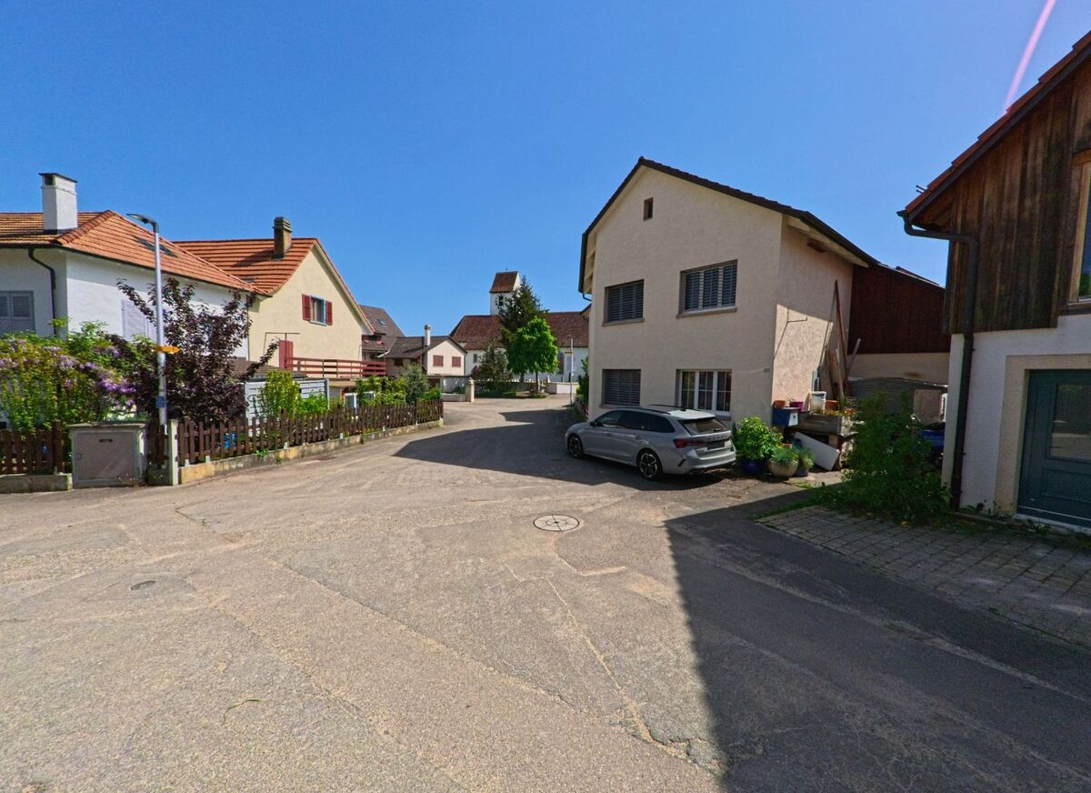 In Nuglar-St. Pantaleon (4421, Kanton Solothurn) fuehrt eine ruhige Quartierstrasse zwischen Einfamilienhaeusern zur Kirche im Hintergrund.