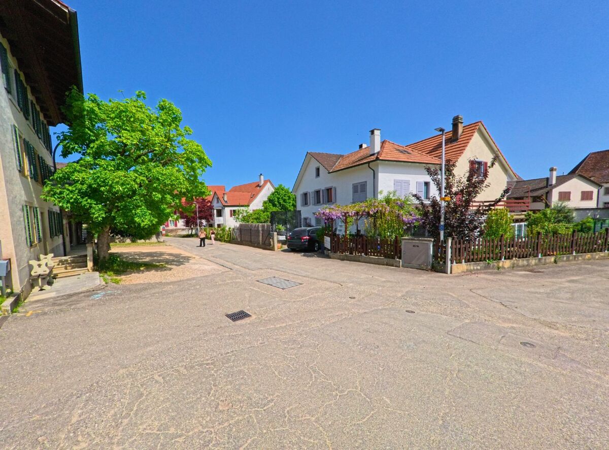 Eine kleine Strasse in Nuglar-St. Pantaleon (4421, Kanton Solothurn) verlaeuft zwischen Wohnhaeusern mit Gaerten, waehrend Passanten im Sonnenschein spazieren.