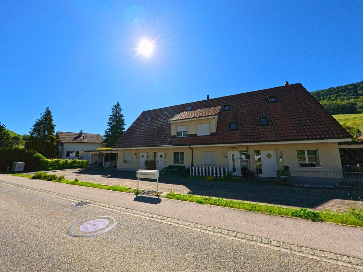 In Nunningen (4208 im Kanton Solothurn) steht ein Mehrfamilienhaus mit rotem Satteldach an einer ruhigen Strasse unter strahlender Sonne.