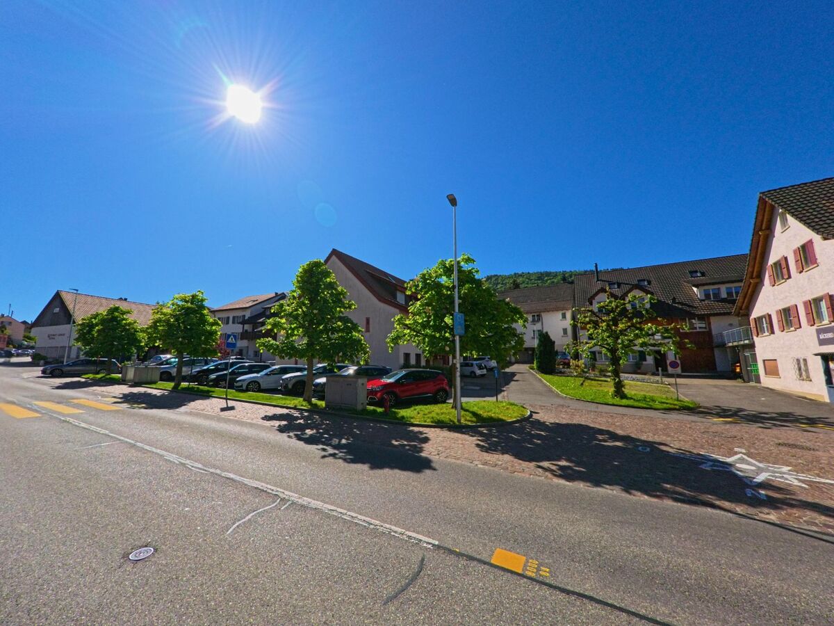 Im Ortszentrum von Nunningen (4208, Kanton Solothurn) säumen junge Bäume einen Parkplatz entlang einer Hauptstrasse bei klarem Sonnenschein.