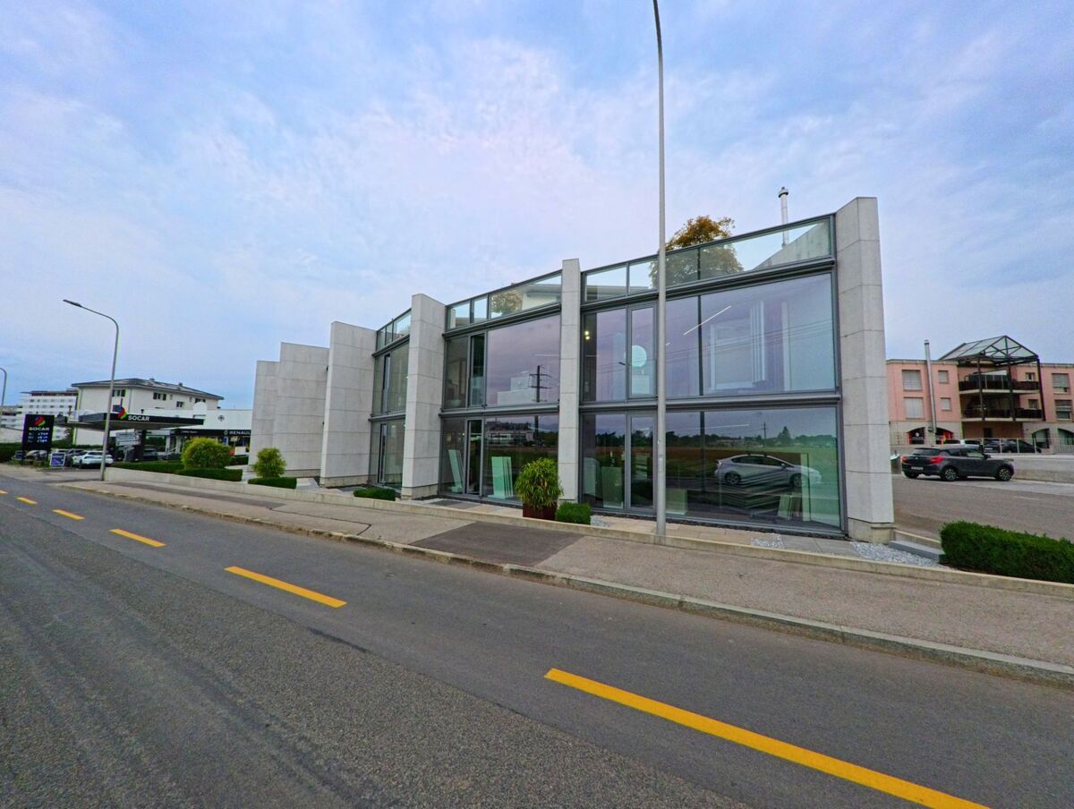 Ein modernes Gebaeude mit grossen Glasfassaden steht in Nyon (1260, Kanton Waadt) direkt an einer Strasse, neben einer Tankstelle und weiteren Wohnhaeusern.