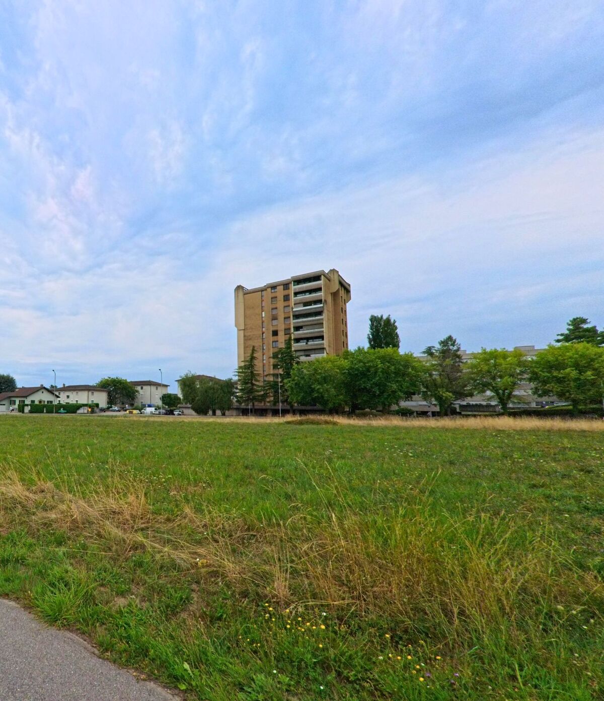 Ein hohes Wohnhaus erhebt sich in Nyon (1260, Kanton Waadt) hinter einer offenen Wiese, umgeben von kleineren Gebaeuden und Baumen am Stadtrand.