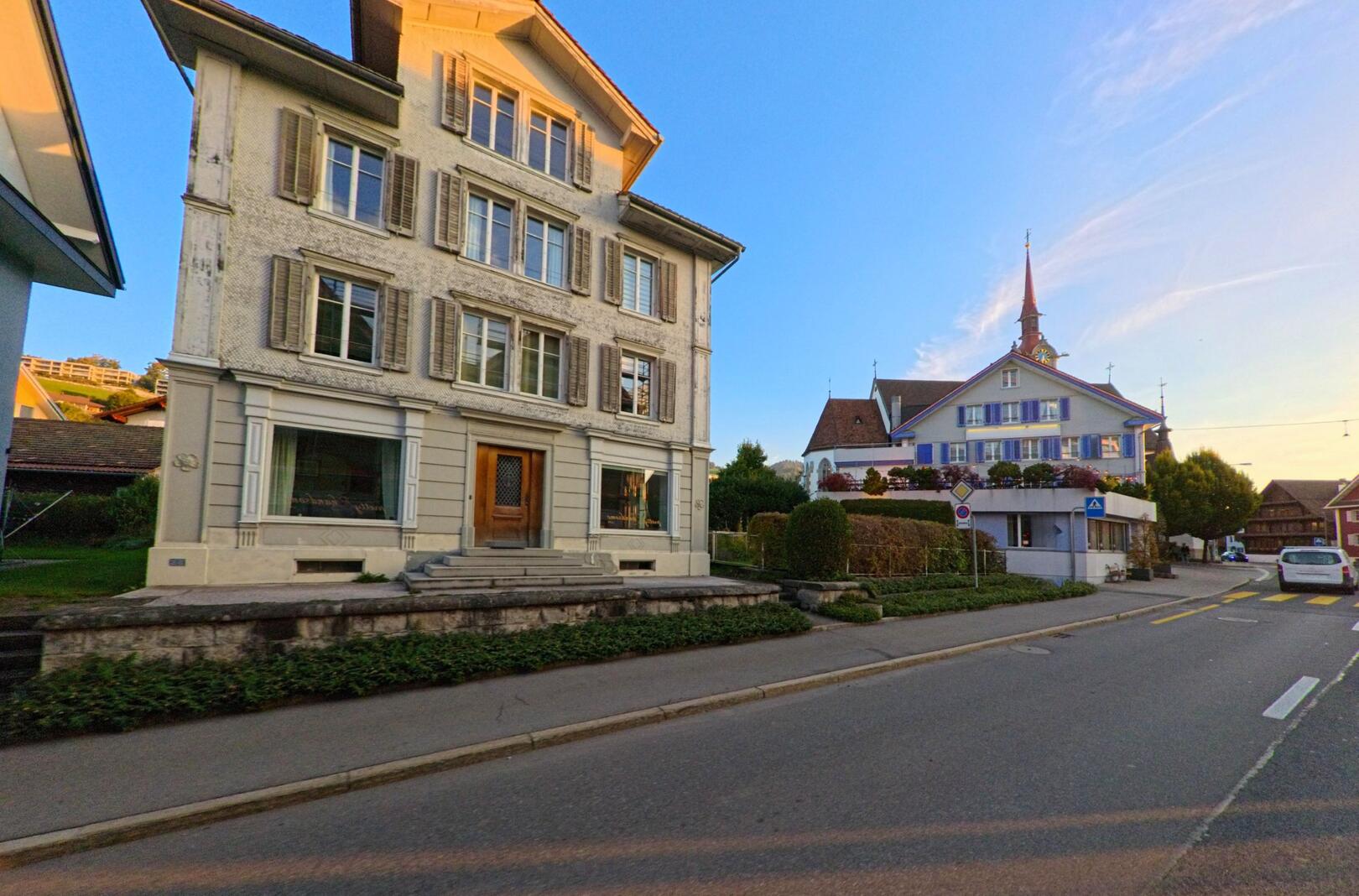 In einer ruhigen Strassenszene in Oberaegeri (6315, Kanton Zug) stehen ein traditionelles Wohnhaus und das Gebaeude mit dem markanten Turm im warmen Abendlicht.