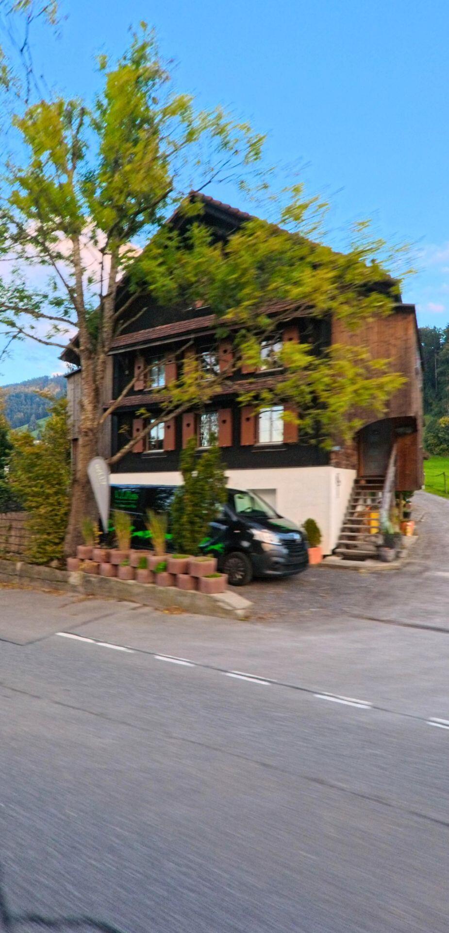 Ein traditionelles Holzhaus in Oberaegeri (6315, Kanton Zug) steht hinter einem grossen Baum, vor dem ein dunkler Lieferwagen auf dem Vorplatz parkiert ist.