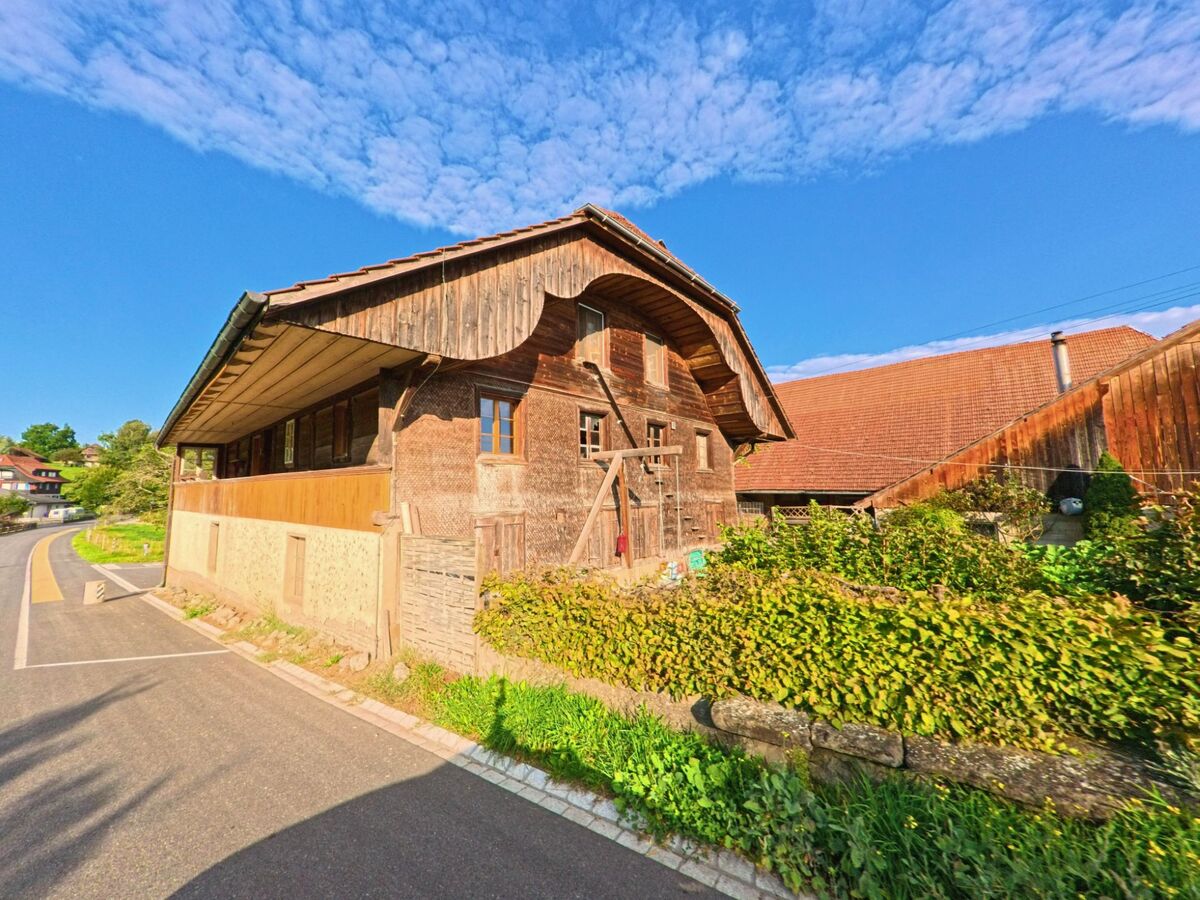 Ein älteres Bauernhaus in Oberbalm (3096, Kanton Bern) steht direkt an einer schmalen Strasse, umgeben von einer niedrigen Steinmauer mit dichter Bepflanzung.