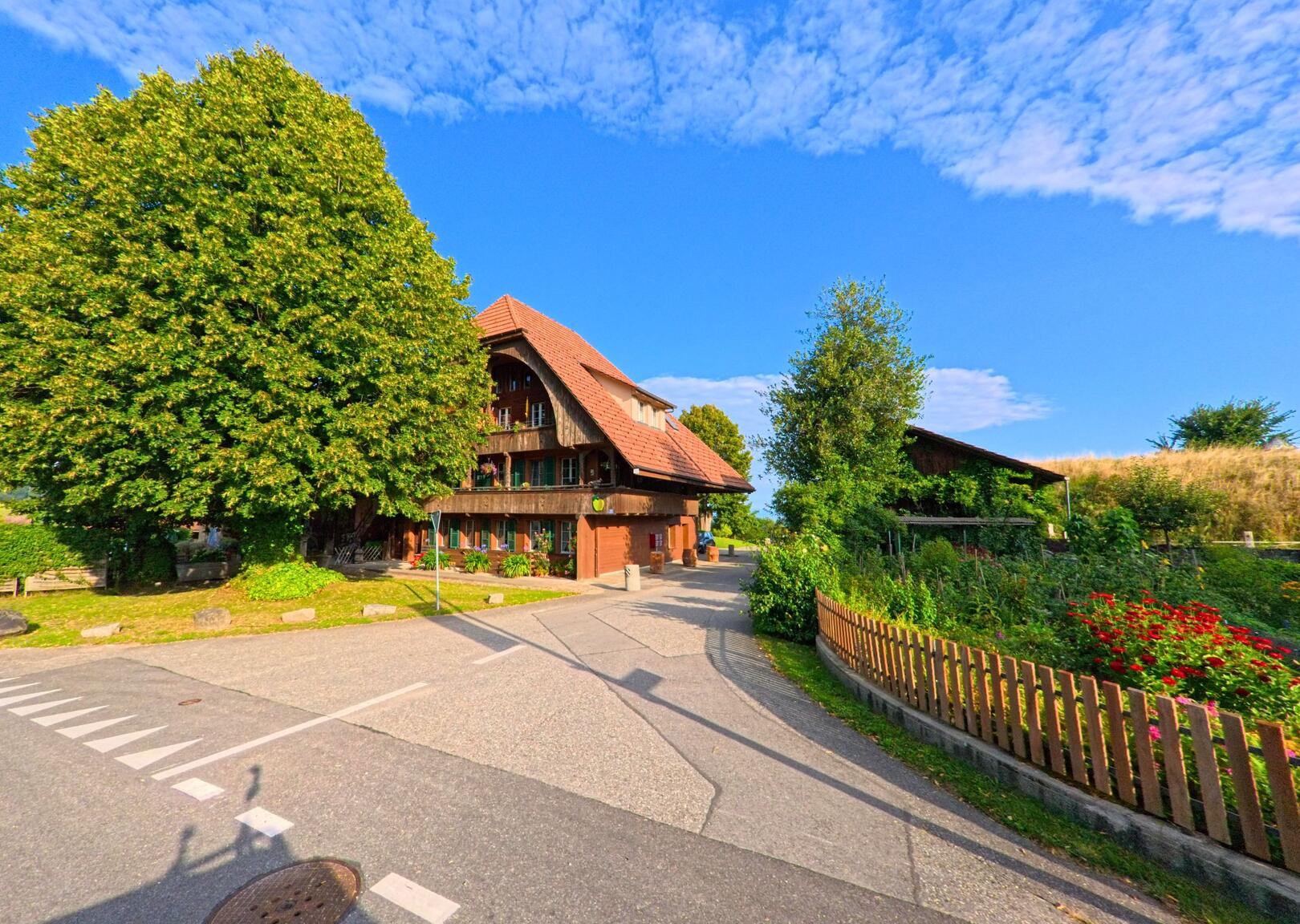 Ein traditionelles Bauernhaus in Oberbalm (3096, Kanton Bern) steht zwischen grossen Laubbäumen, umgeben von einem Garten und angrenzenden Wirtschaftsgebäuden unter blauem Himmel.