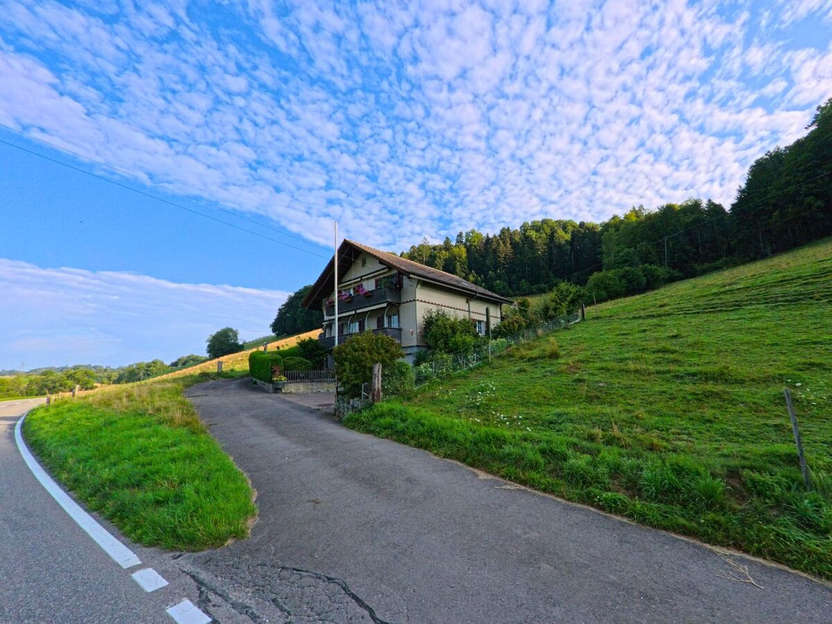 Ein Einfamilienhaus in Oberbalm (3096, Kanton Bern) liegt am Rand einer Wiese, mit direktem Blick auf den angrenzenden Waldrand unter leicht bewölktem Himmel.