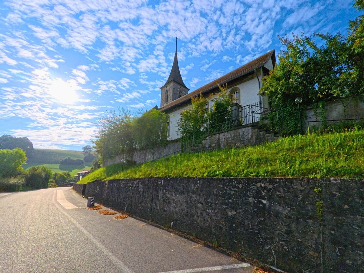 Die Kirche von Oberbalm (3096, Kanton Bern) steht erhöht auf einer Mauerterrasse, umgeben von Bäumen und mit Blick auf die sanfte Hügellandschaft im Hintergrund.