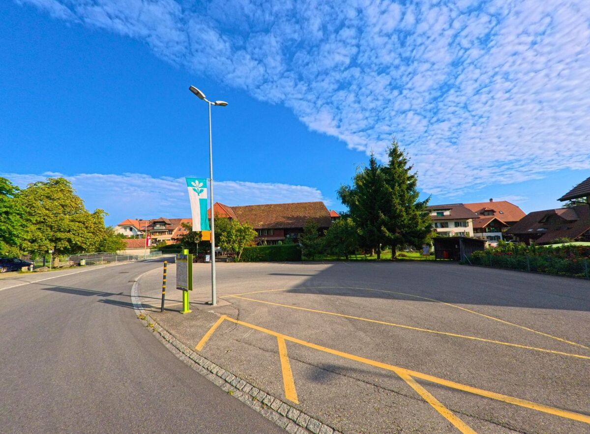 Ein öffentlicher Platz in Oberbalm (3096, Kanton Bern) mit markierten Parkflächen, umgeben von Wohnhäusern und Bäumen unter klarem Sommerhimmel.