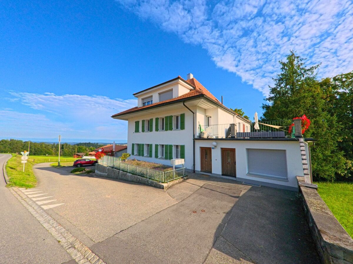Ein helles Wohnhaus mit grünen Fensterläden in Oberbalm (3096, Kanton Bern) steht an einer leicht kurvigen Strasse, umgeben von Wiesen und mit weitem Blick in die Landschaft.