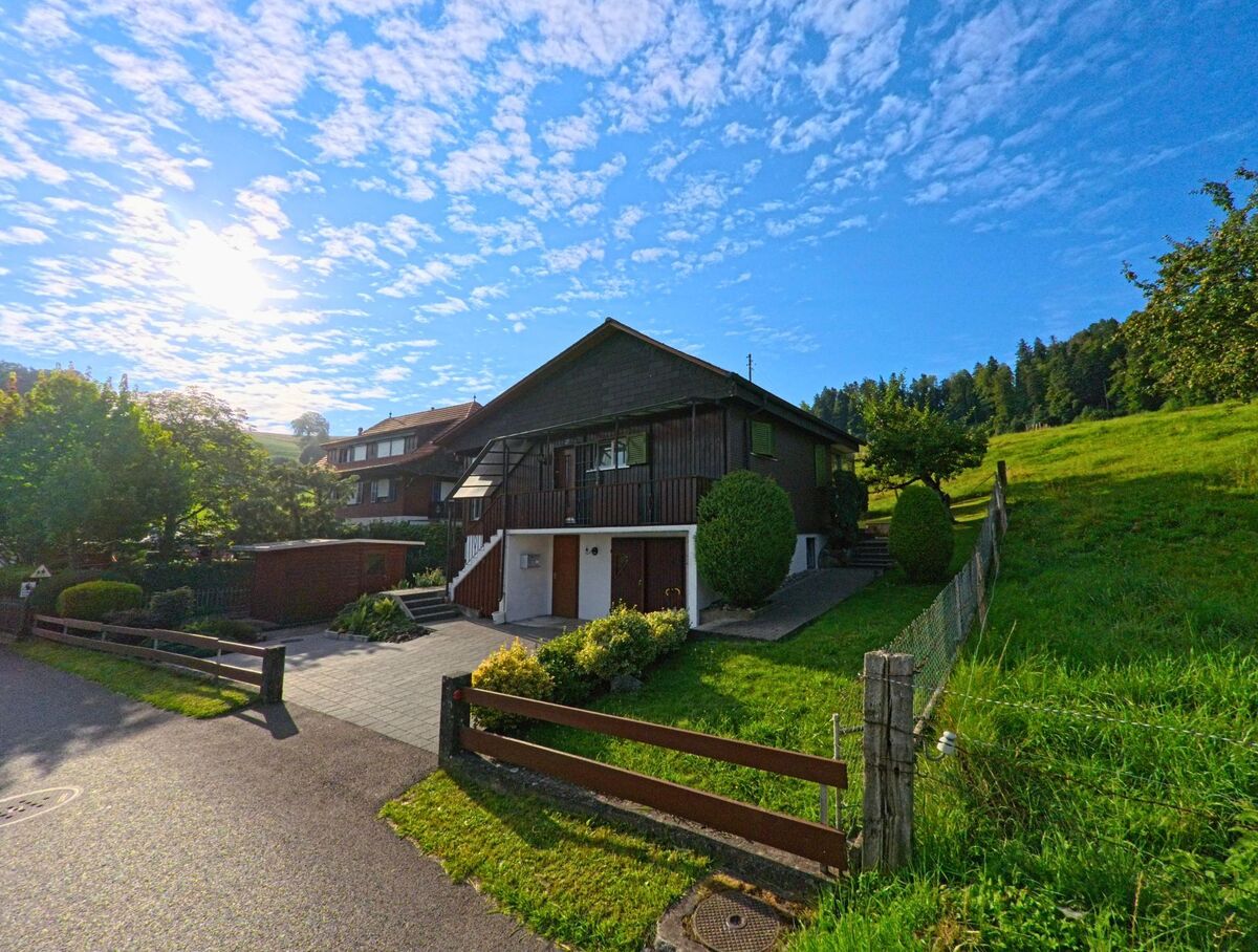 Ein Wohnhaus mit dunkler Holzfassade in Oberbalm (3096, Kanton Bern) steht am Rand einer Wiese, umgeben von gepflegten Büschen und einem Gartenzaun.