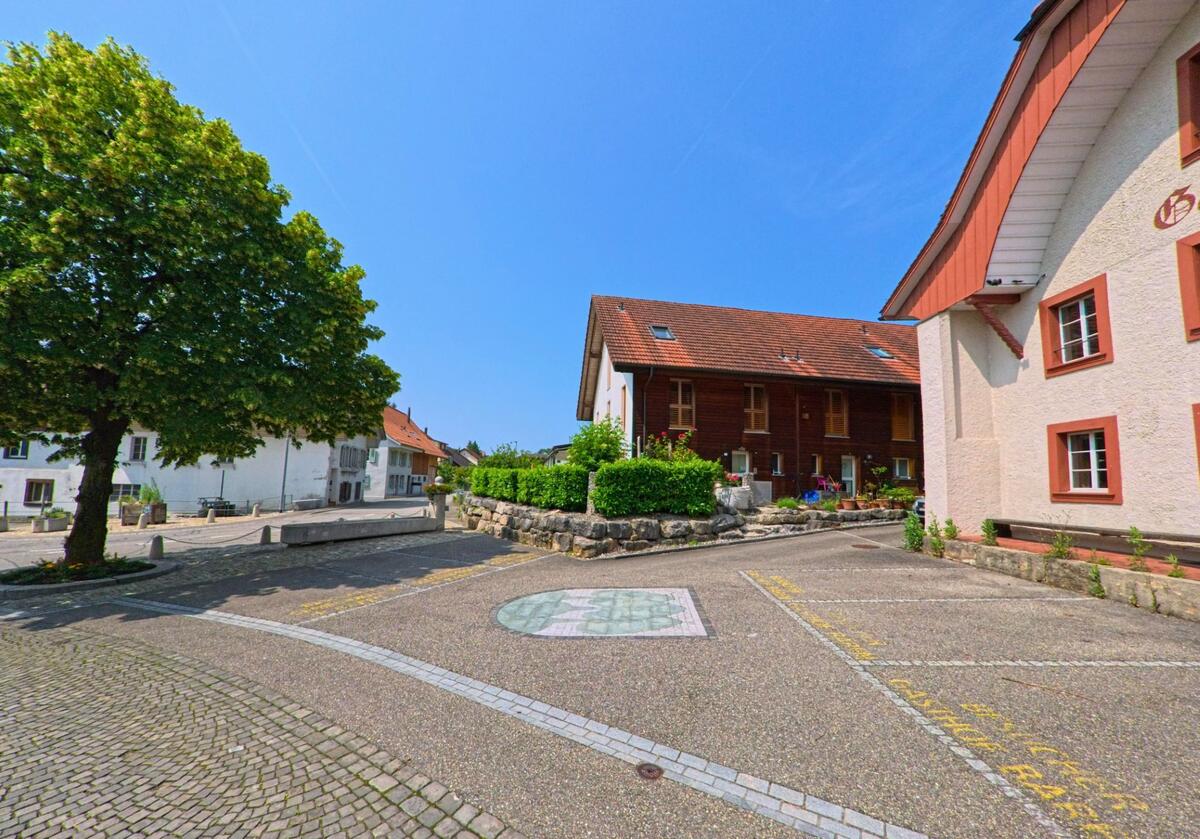 In Oberbipp (4538, Kanton Bern) liegt ein sonniger Platz mit Kopfsteinpflaster, einem schattenspendenden Baum und angrenzenden Wohnhäusern mit gepflegten Gärten.