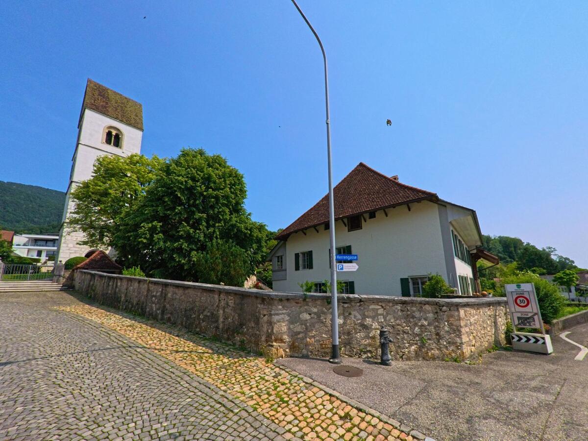 In der Herrengasse von Oberbipp (4538, Kanton Bern) steht ein historisches Wohnhaus mit grünen Fensterläden gegenüber dem Kirchturm, umgeben von einer Steinmauer und alten Pflastersteinen.
