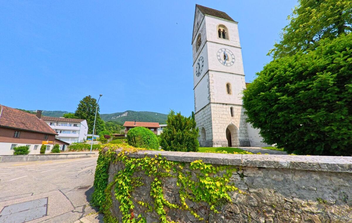 Der markante Uhrturm der reformierten Kirche in Oberbipp (4538, Kanton Bern) ragt über eine mit Efeu bewachsene Steinmauer vor dem Hintergrund der Jurahügel hinaus.