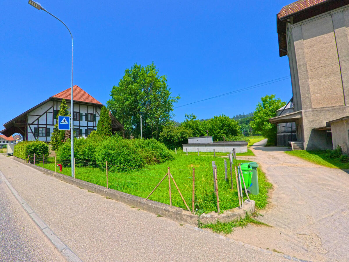 In Oberburg (3414, Kanton Bern) liegt ein gepflegtes Fachwerkhaus mit Vorgarten gegenüber einem grossen Betongebäude, verbunden durch einen schmalen Weg entlang einer Grünfläche.