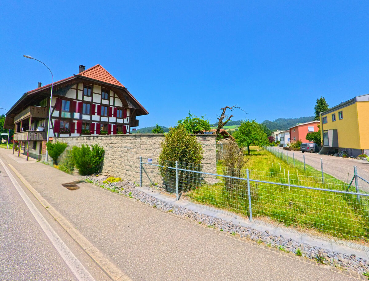 In Oberburg (3414, Kanton Bern) steht ein traditionelles Bauernhaus mit rotem Ziegeldach und Fachwerkfassade neben einer Mauer und modernen Wohnbauten.