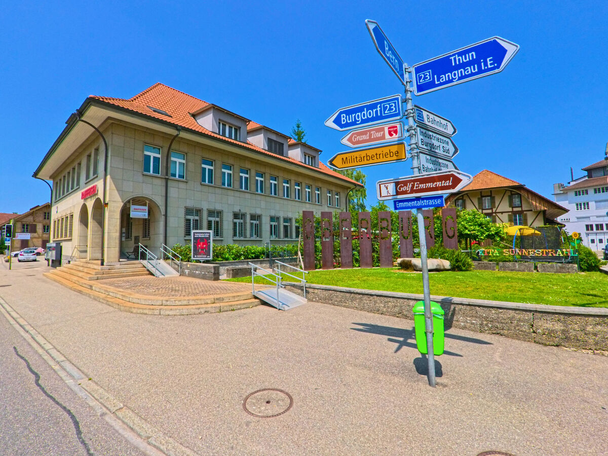 Im Zentrum von Oberburg (3414, Kanton Bern) steht das markante Gemeindehaus mit Treppenaufgang, flankiert von Wegweisern und dem Schriftzug „Oberburg“ im Grünbereich.